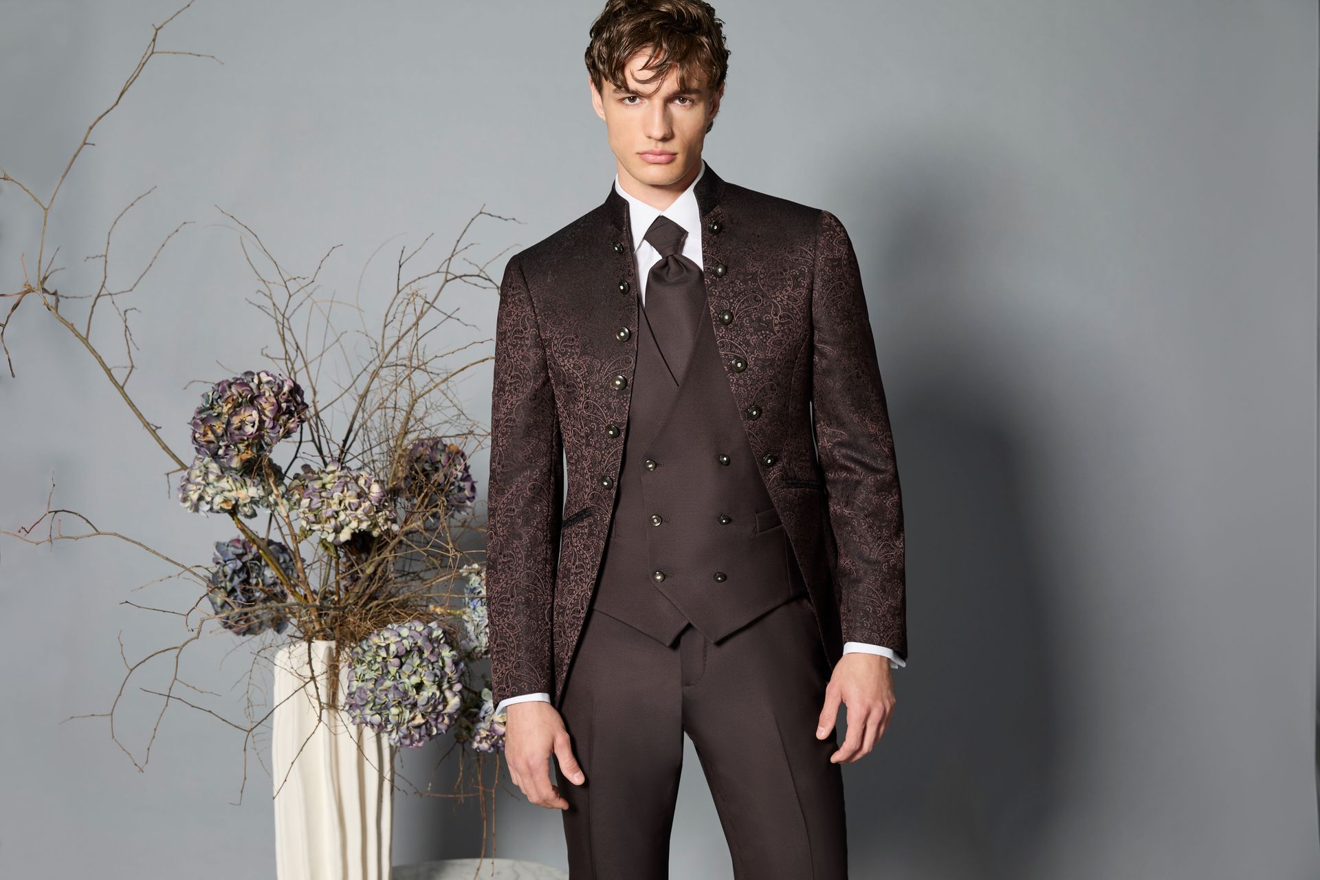 Uomo in abito e gilet marrone scuro con fantasia, camicia bianca e cravatta; in piedi vicino ai fiori.