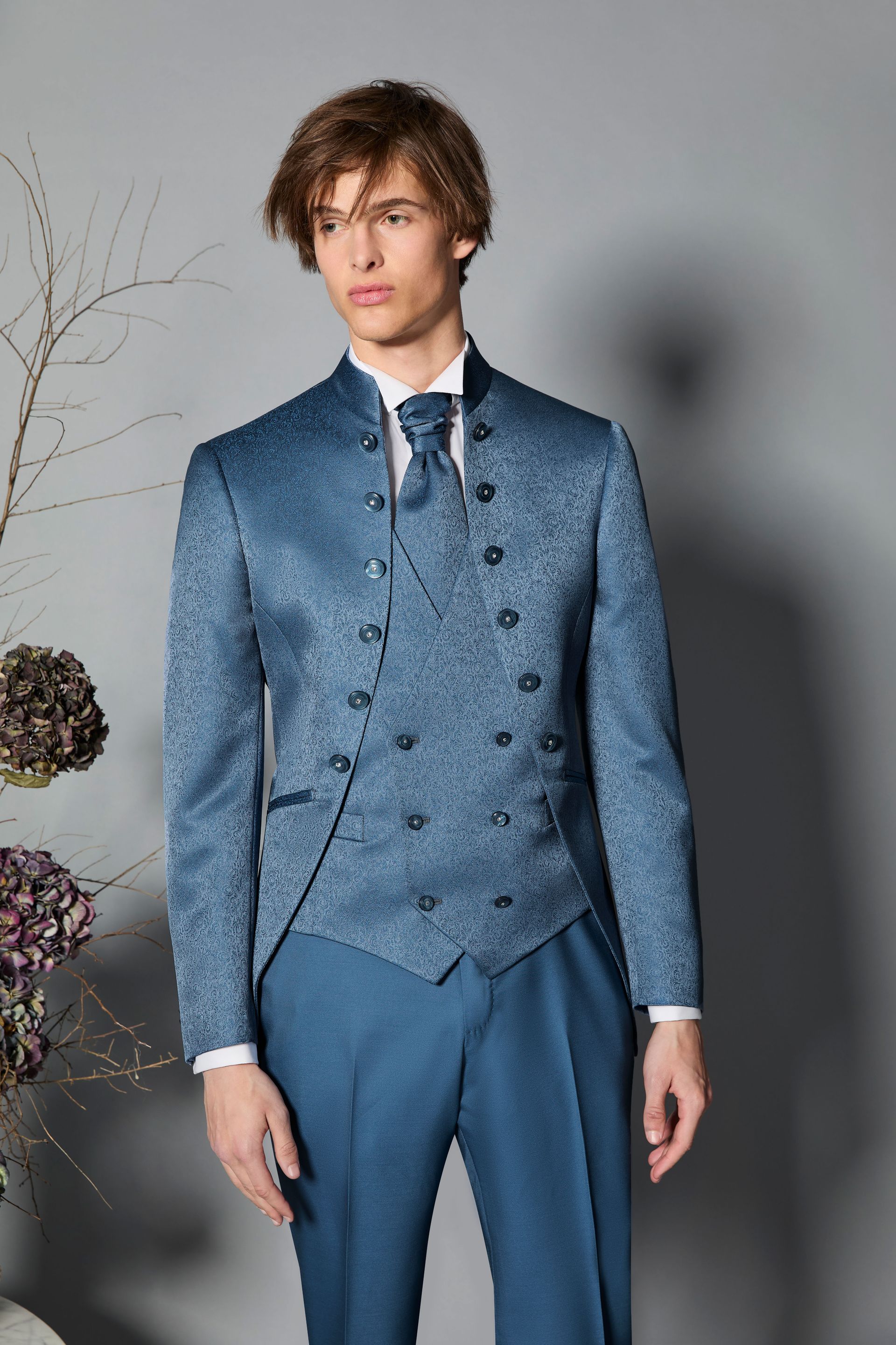 Uomo in abito e gilet blu fantasia, camicia bianca e cravatta, in posa in un interno.