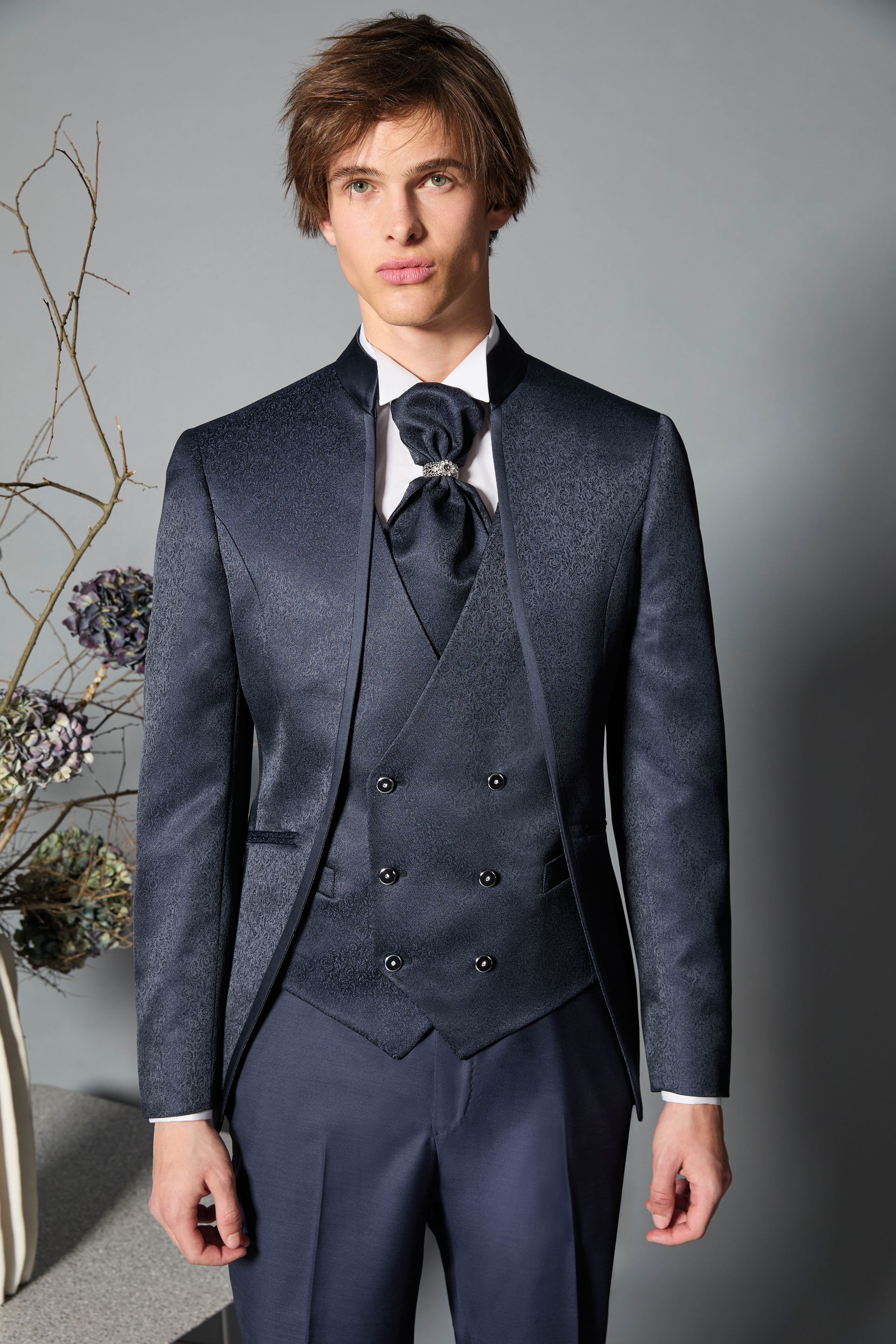 Uomo in un elegante abito blu con giacca a fantasia, gilet e cravatta, in piedi in un ambiente interno.