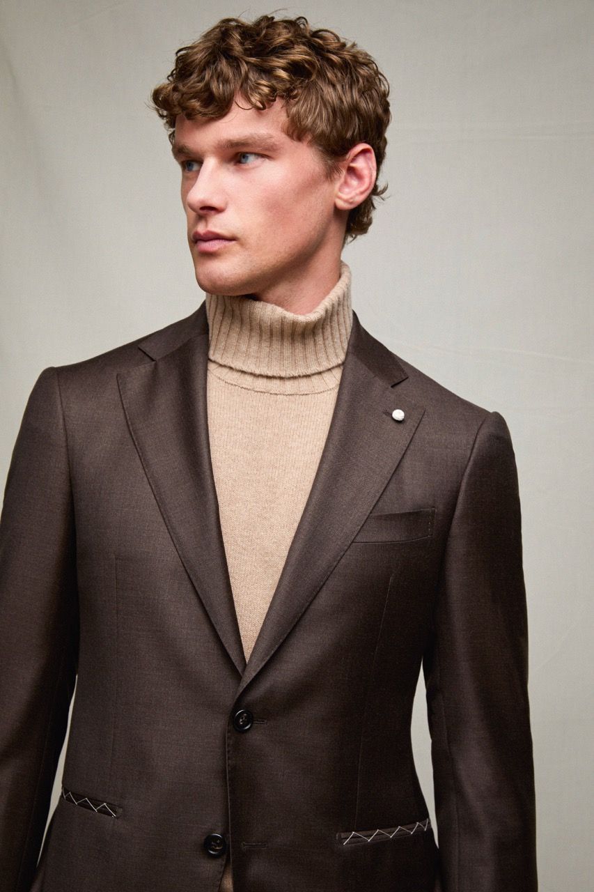 Uomo con blazer marrone e dolcevita beige, che guarda di lato su uno sfondo grigio uniforme