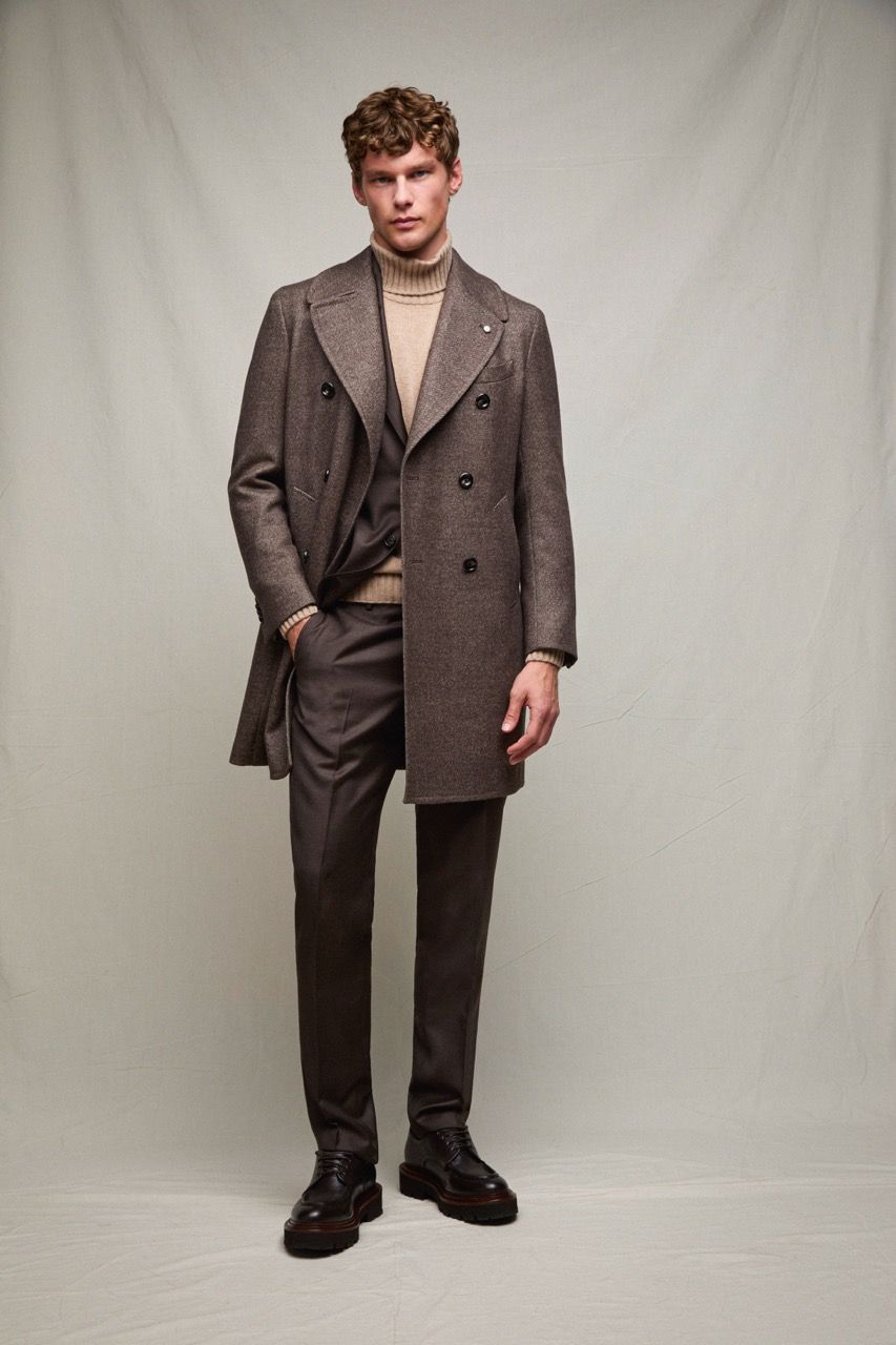 Uomo in piedi su uno sfondo grigio da studio, con un abito marrone e un dolcevita beige.