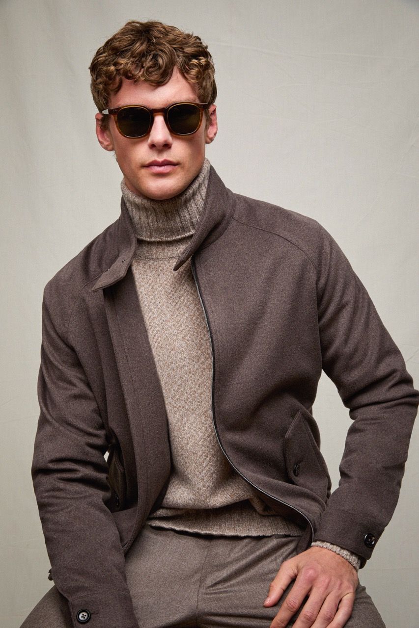 Uomo con occhiali da sole, giacca marrone e dolcevita grigio, seduto davanti a uno sfondo chiaro e uniforme.
