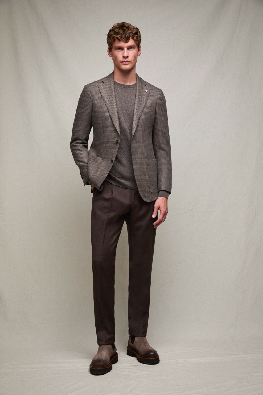 Uomo con blazer grigio, pantaloni scuri e stivali, in piedi davanti a uno sfondo neutro.