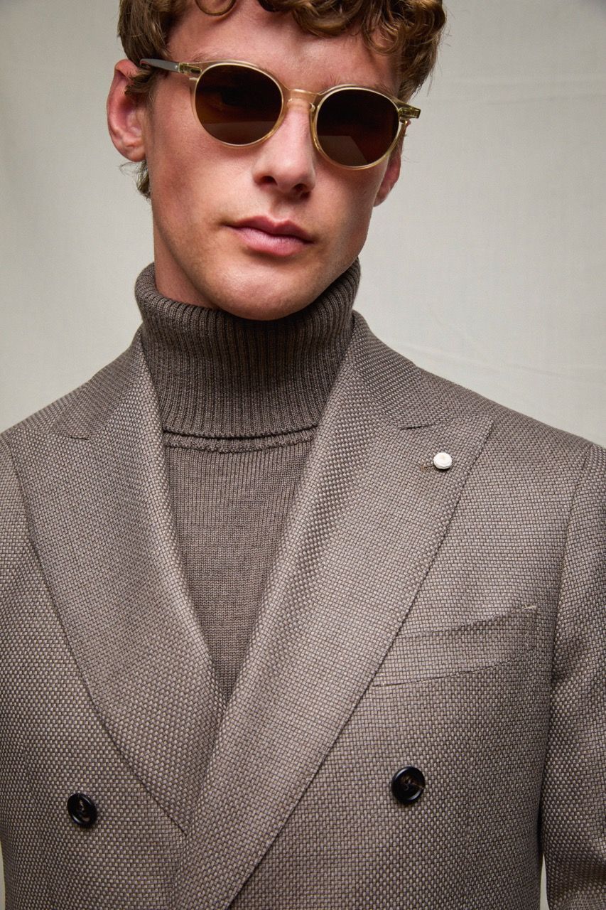 Persona con occhiali da sole rotondi e blazer grigio a spina di pesce sopra un dolcevita.