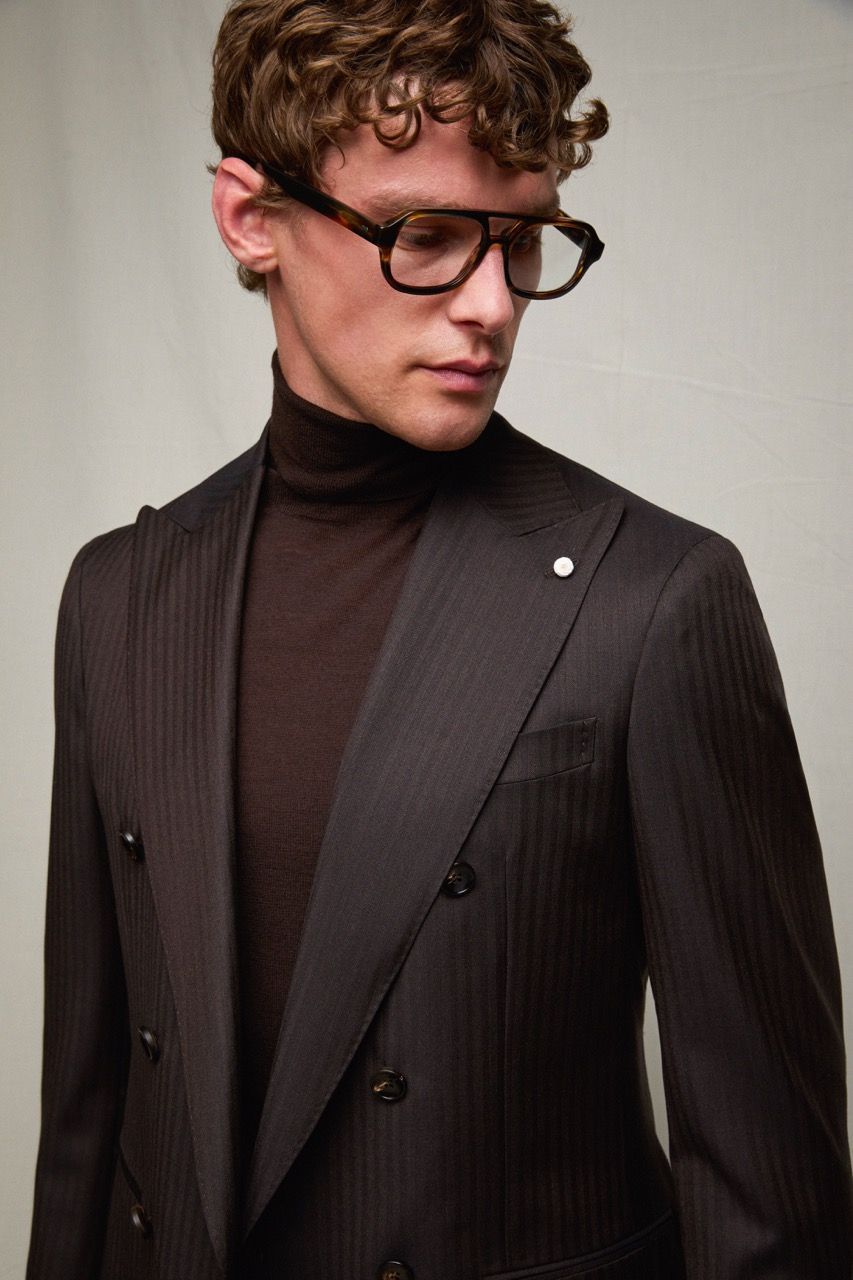 Uomo con blazer scuro a righe sottili sopra un dolcevita marrone, con gli occhiali, che guarda in basso.