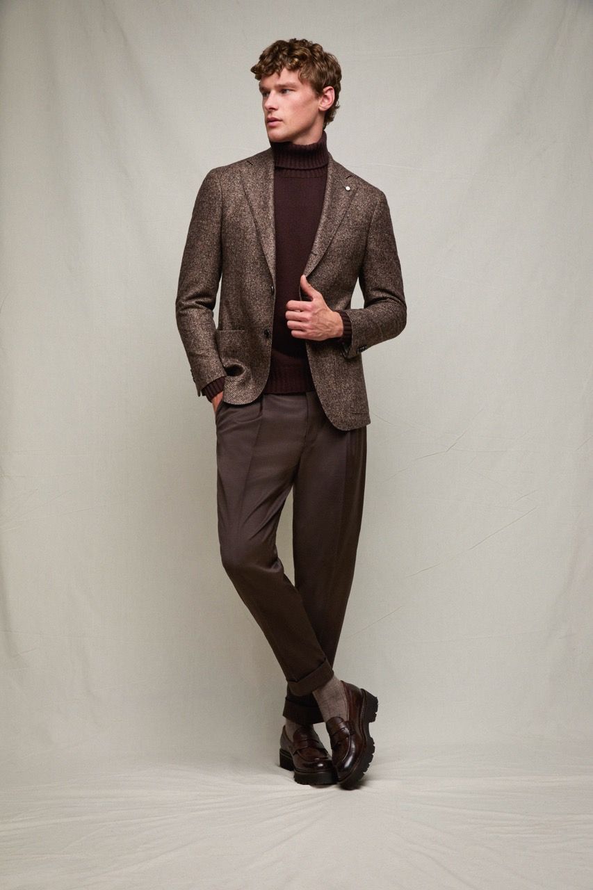 Uomo in posa con blazer marrone fantasia, dolcevita e pantaloni, davanti a uno sfondo neutro da studio