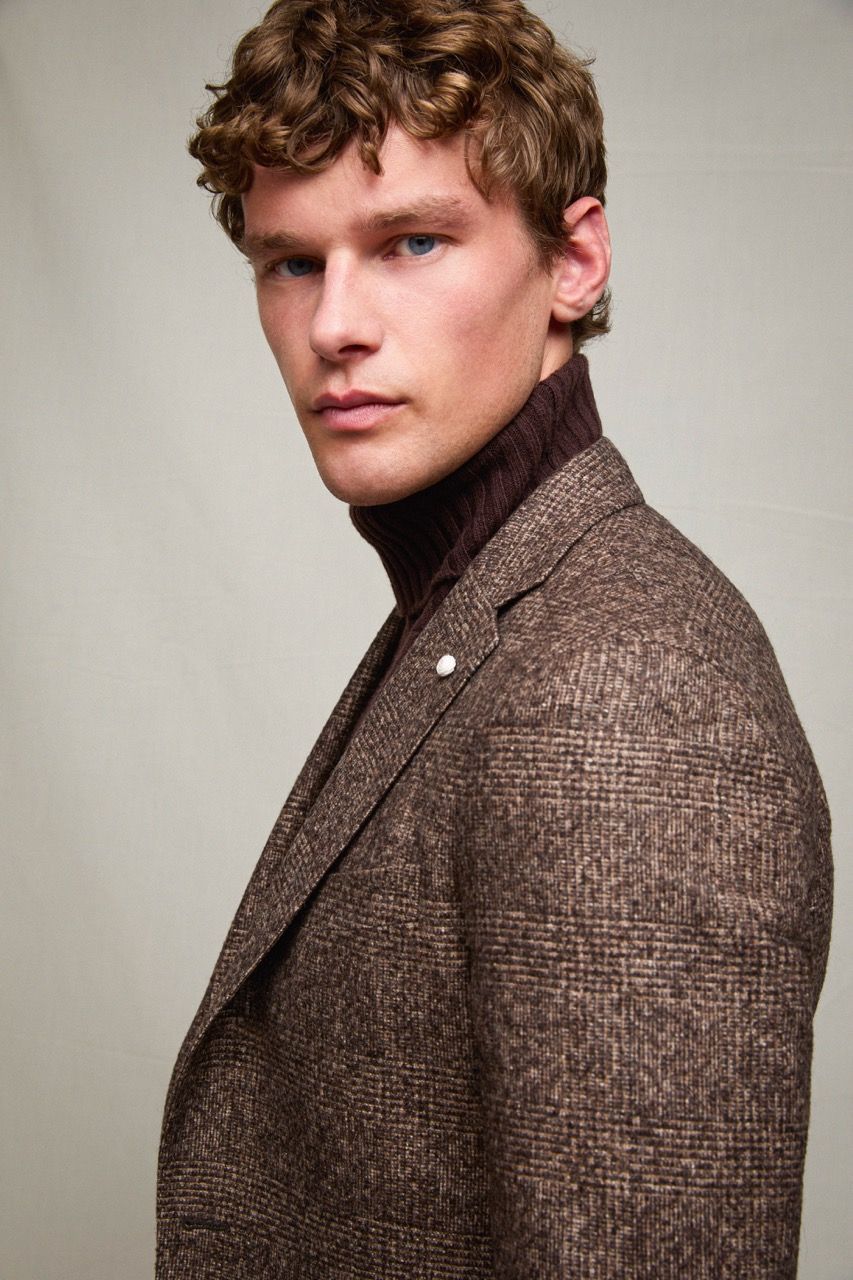 Ritratto di un uomo con blazer in tweed marrone e dolcevita scuro, che guarda al di sopra della spalla su uno sfondo grigio