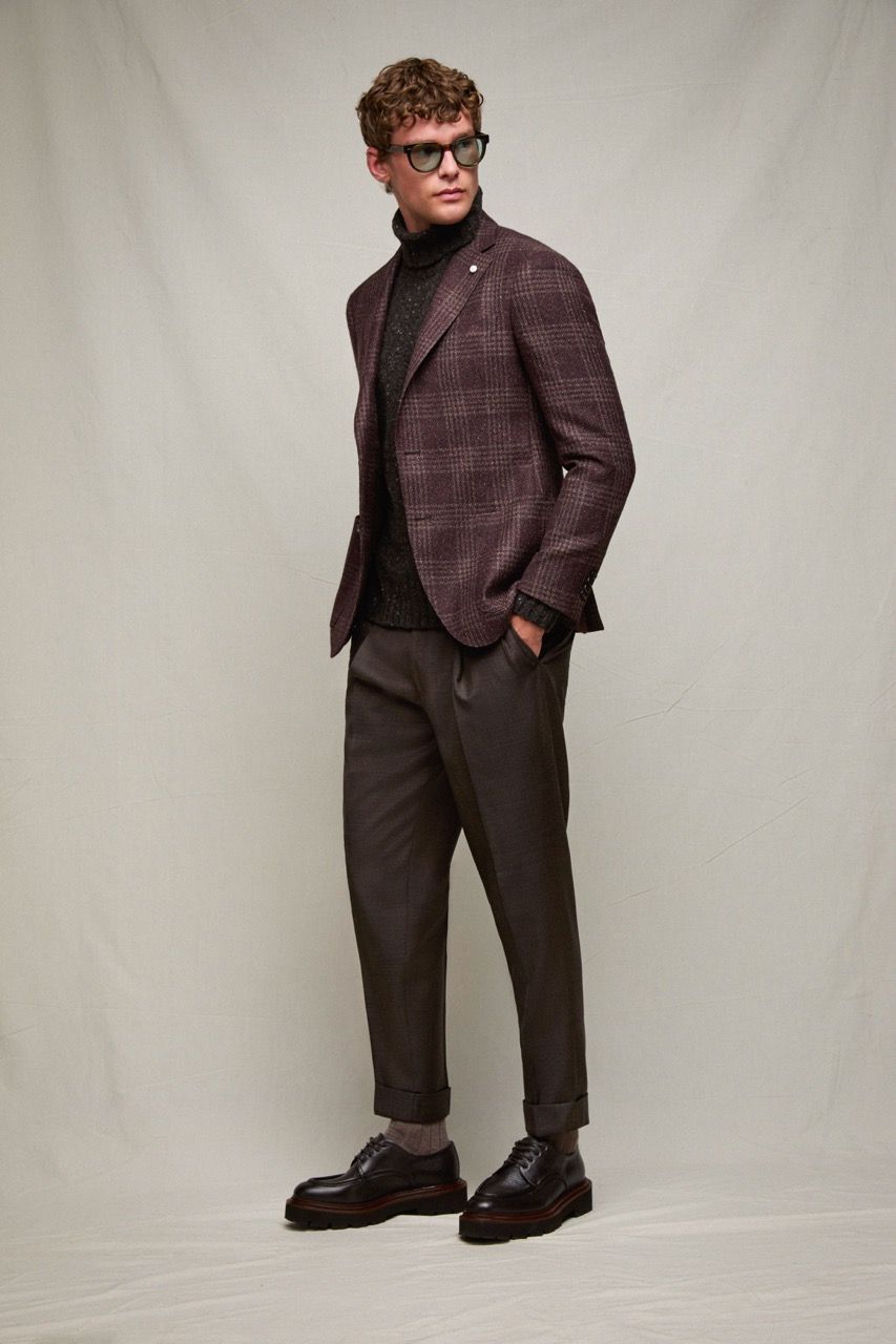 Uomo con blazer color bordeaux e pantaloni scuri, in piedi davanti a uno sfondo neutro e con lo sguardo rivolto di lato.