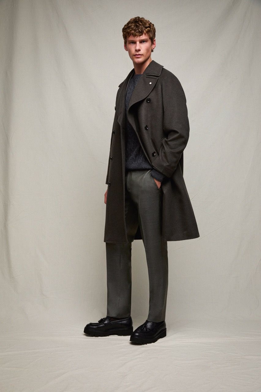 Uomo con cappotto e pantaloni scuri in piedi davanti a uno sfondo grigio uniforme