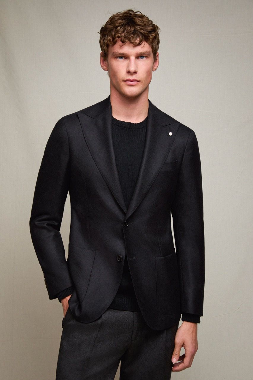 Persona con giacca e camicia nere in piedi davanti a uno sfondo grigio uniforme.
