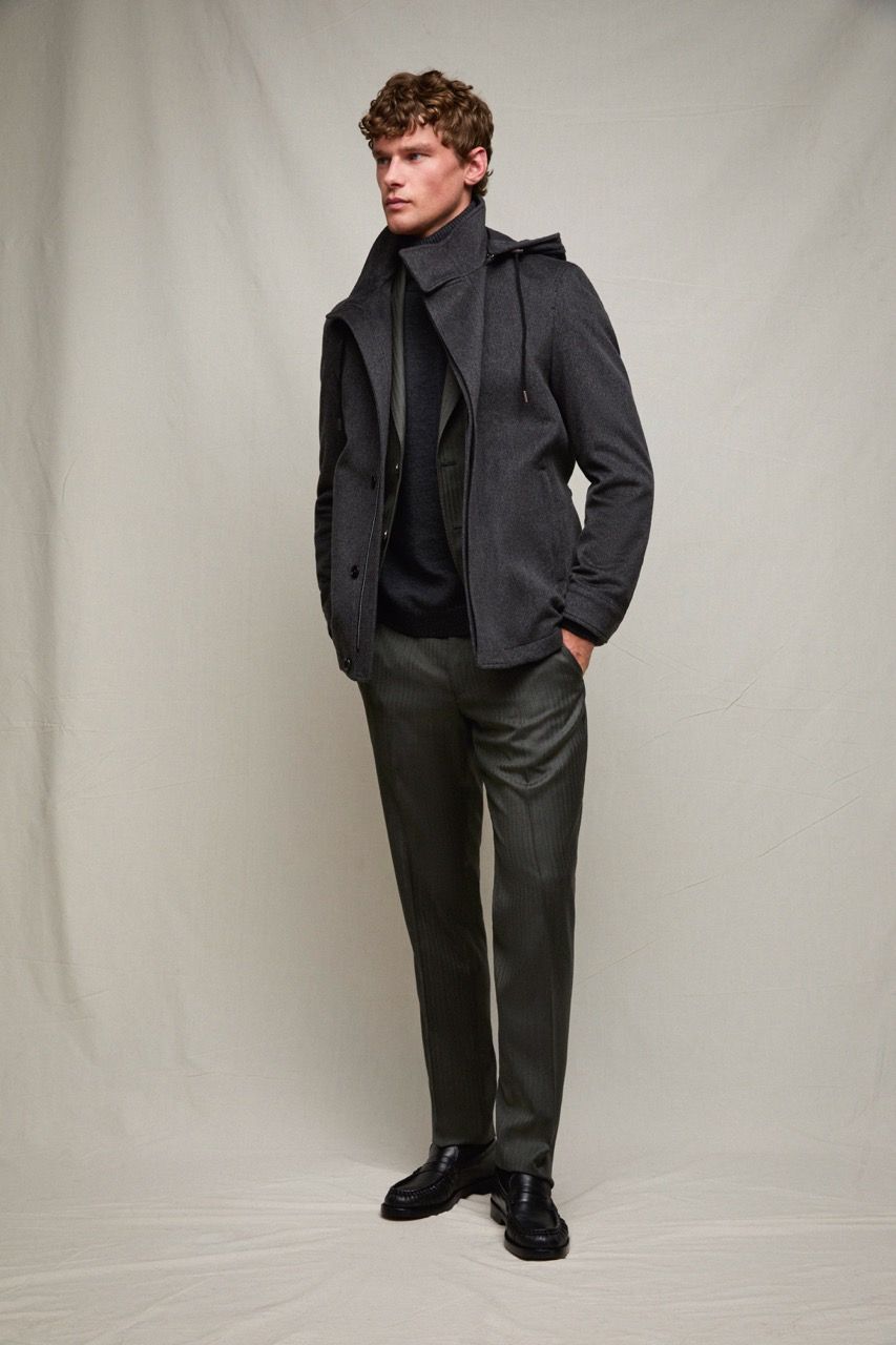 Uomo con giacca e pantaloni scuri in piedi davanti a uno sfondo neutro da studio.
