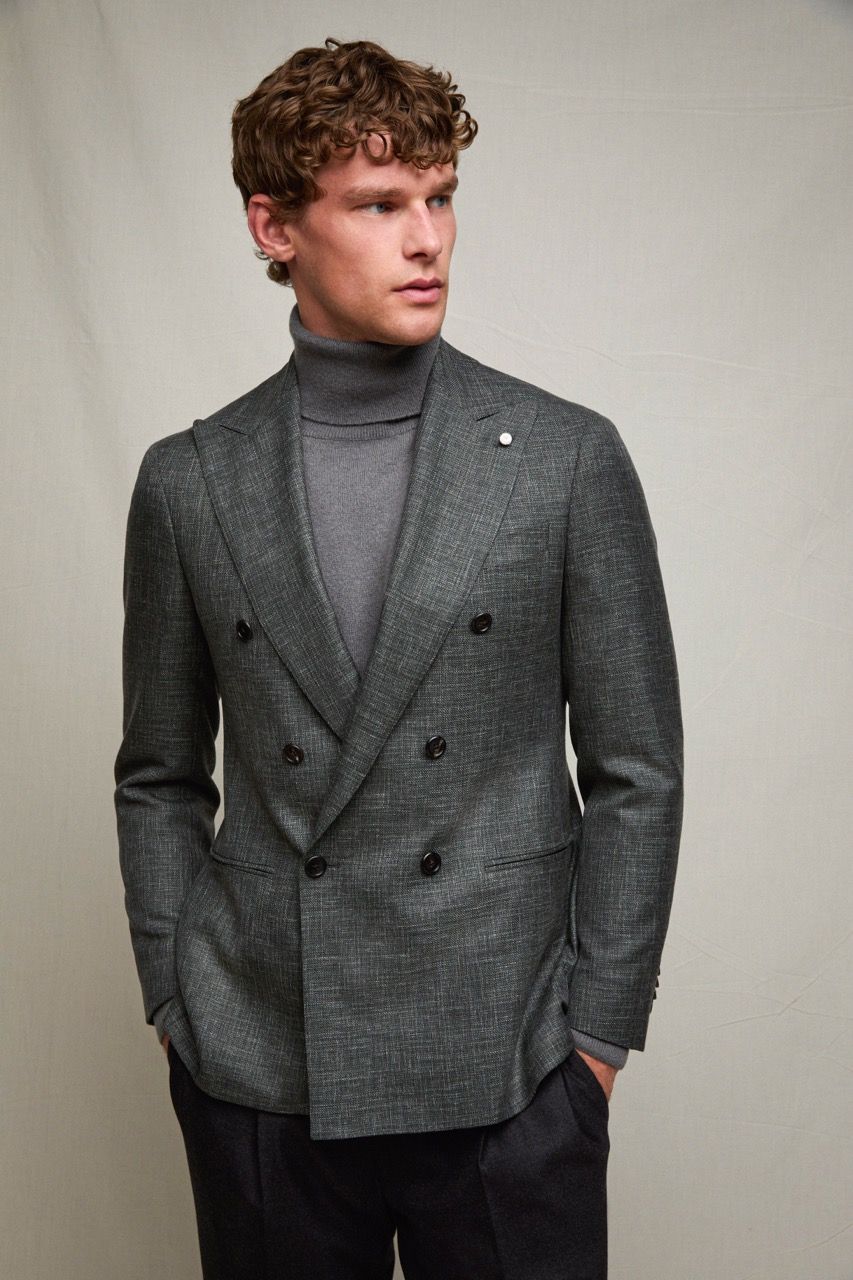 Uomo con blazer doppiopetto grigio e dolcevita, in piedi davanti a uno sfondo neutro e con lo sguardo rivolto a sinistra