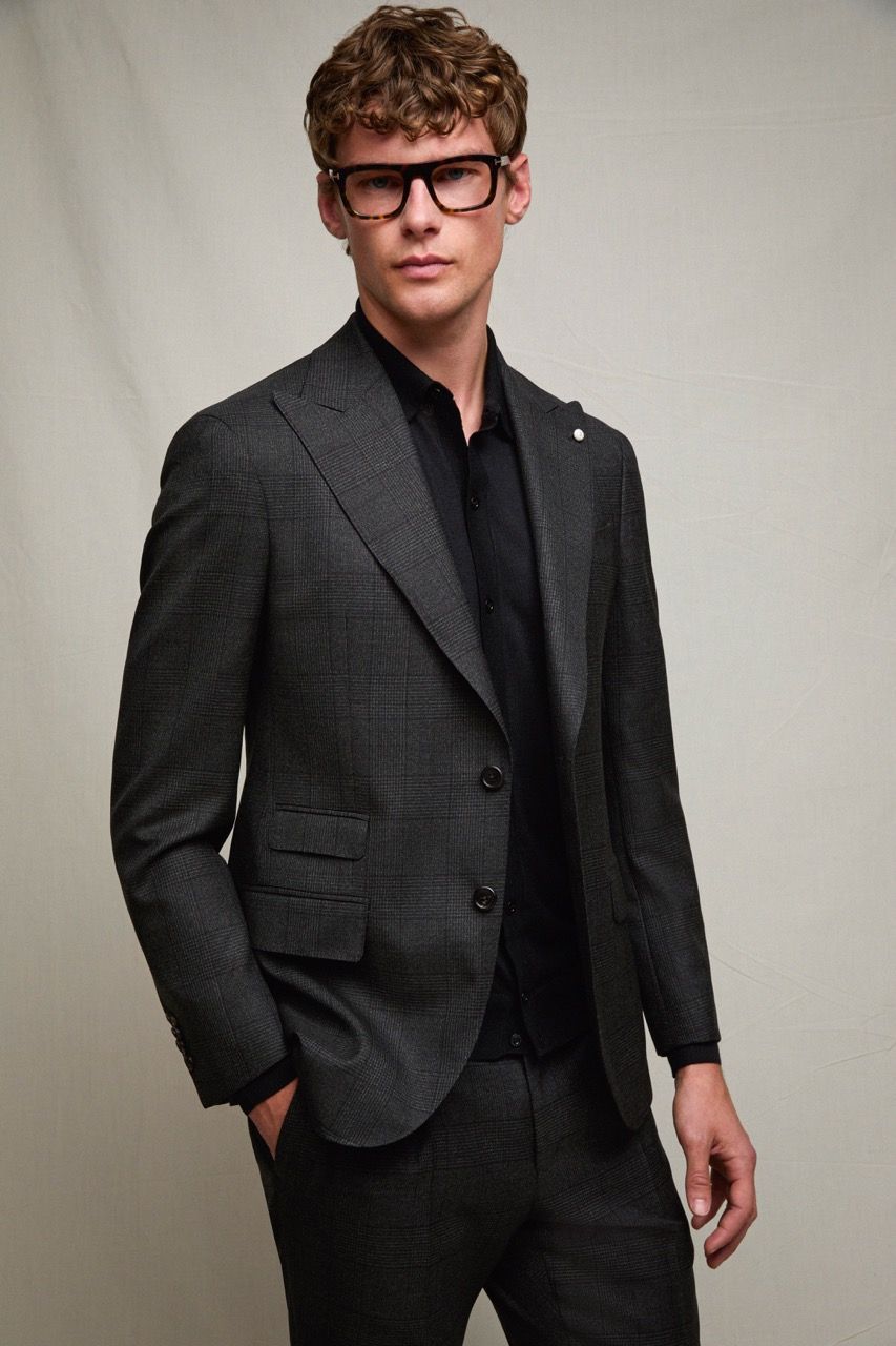 Uomo in abito grigio scuro e camicia nera, in posa contro una parete spoglia, con gli occhiali.
