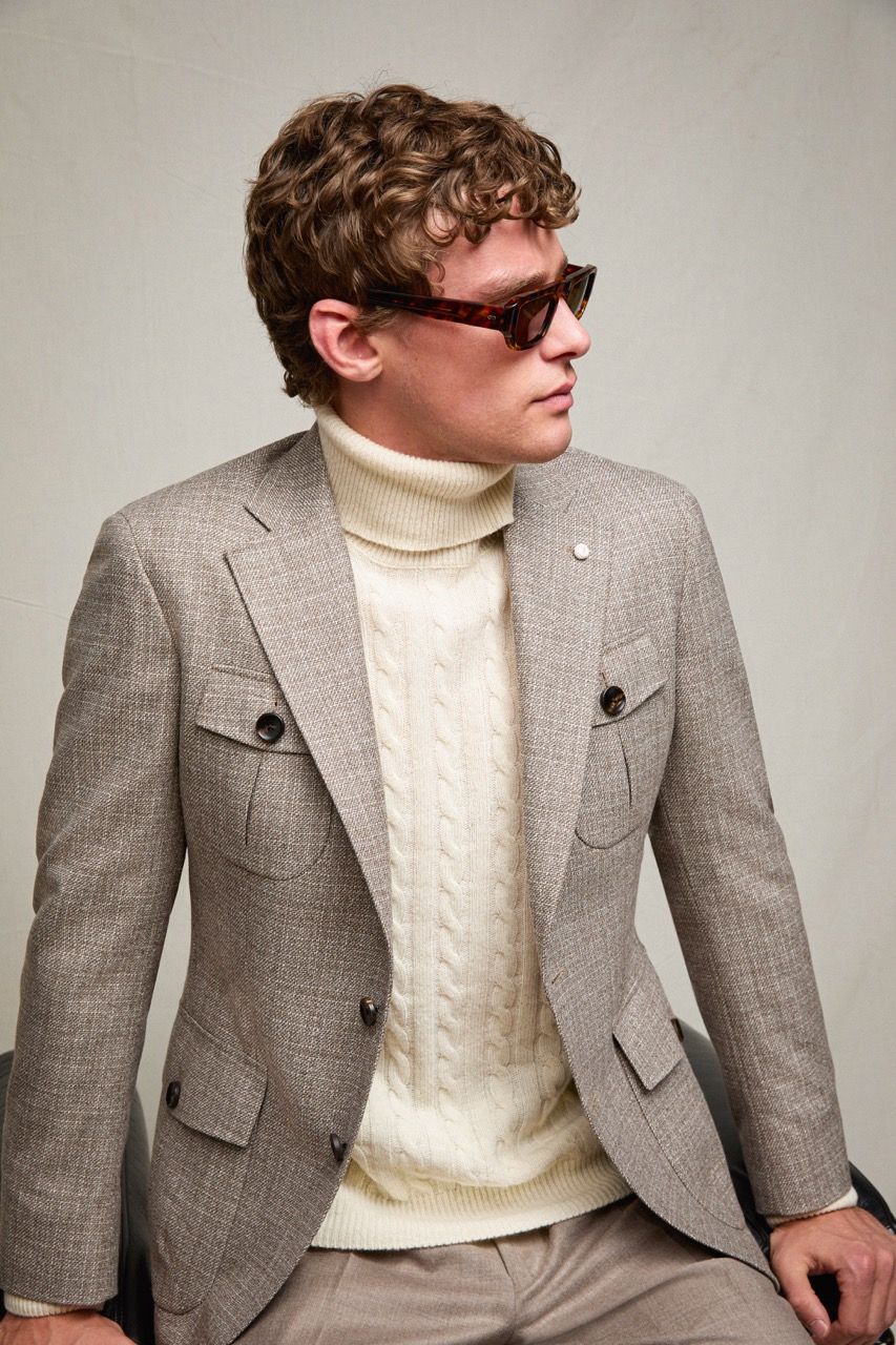 Uomo con blazer grigio e dolcevita color crema, con occhiali da sole, seduto davanti a una parete spoglia