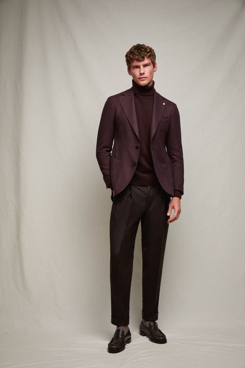 Uomo in piedi con un blazer bordeaux e un dolcevita nero, su uno sfondo neutro.