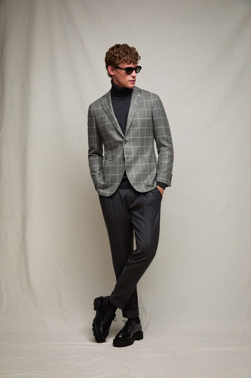 Uomo con blazer grigio e pantaloni neri in posa davanti a uno sfondo neutro, con gli occhiali da sole.