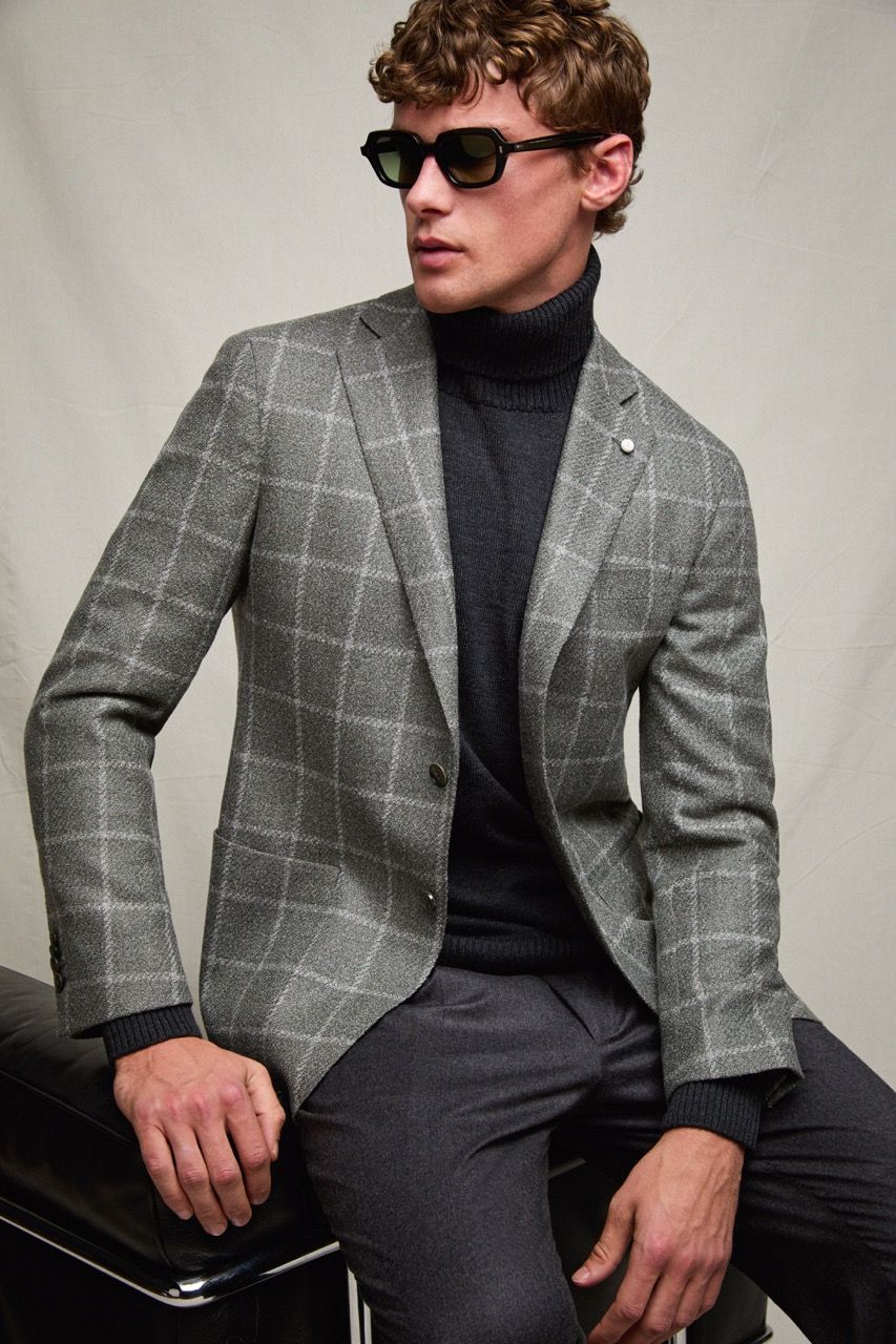Uomo con blazer grigio a quadri e dolcevita nero, seduto davanti a uno sfondo neutro, con gli occhiali da sole.