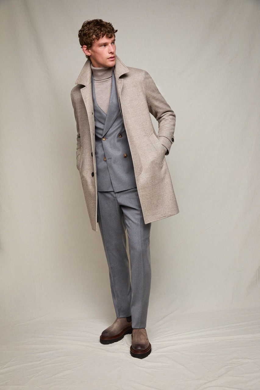 Uomo in abito grigio e cappotto beige in posa davanti a uno sfondo neutro