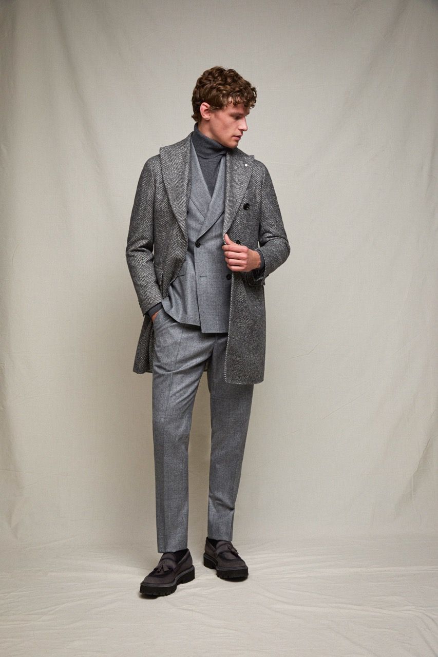 Uomo in abito grigio e camicia fantasia, in piedi davanti a uno sfondo neutro e con lo sguardo rivolto di lato.