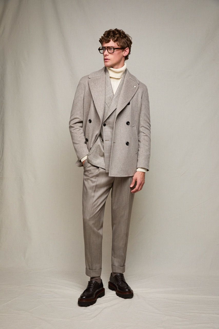 Uomo con un abito doppiopetto grigio chiaro e scarpe nere, in piedi davanti a uno sfondo grigio uniforme.