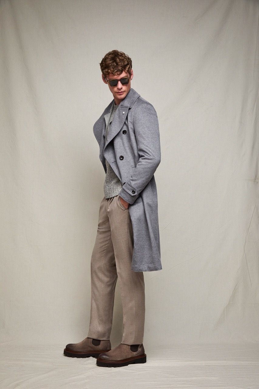 Persona con cappotto grigio, pantaloni beige e scarpe marroni in posa su uno sfondo chiaro.