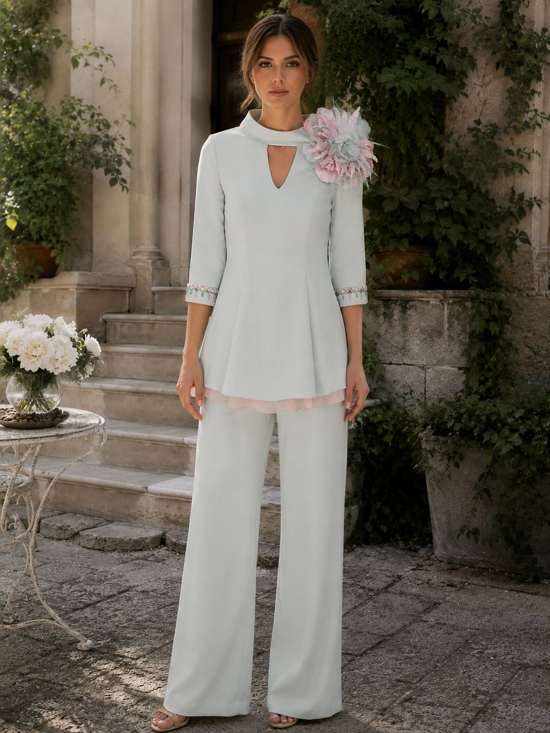Donna con una camicetta bianca e pantaloni a gamba larga in piedi in un cortile con giardino e fiori rosa.
