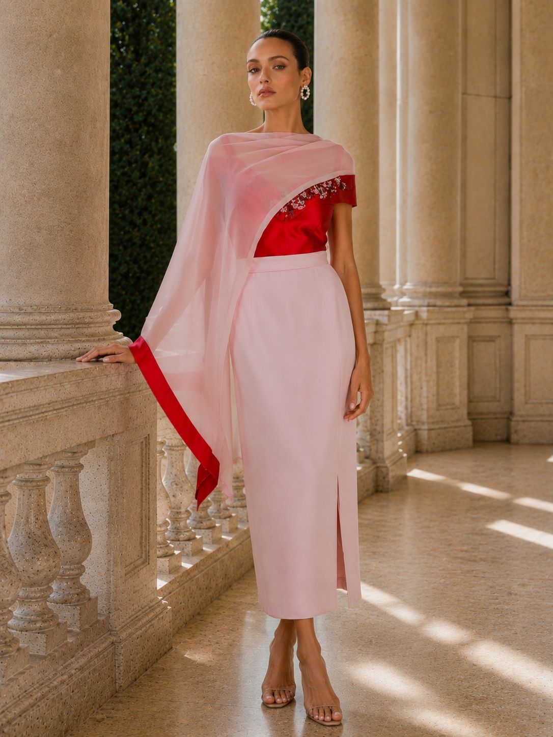Modella con un abito rosa pallido e dettagli rossi, in piedi in un porticato illuminato dal sole.