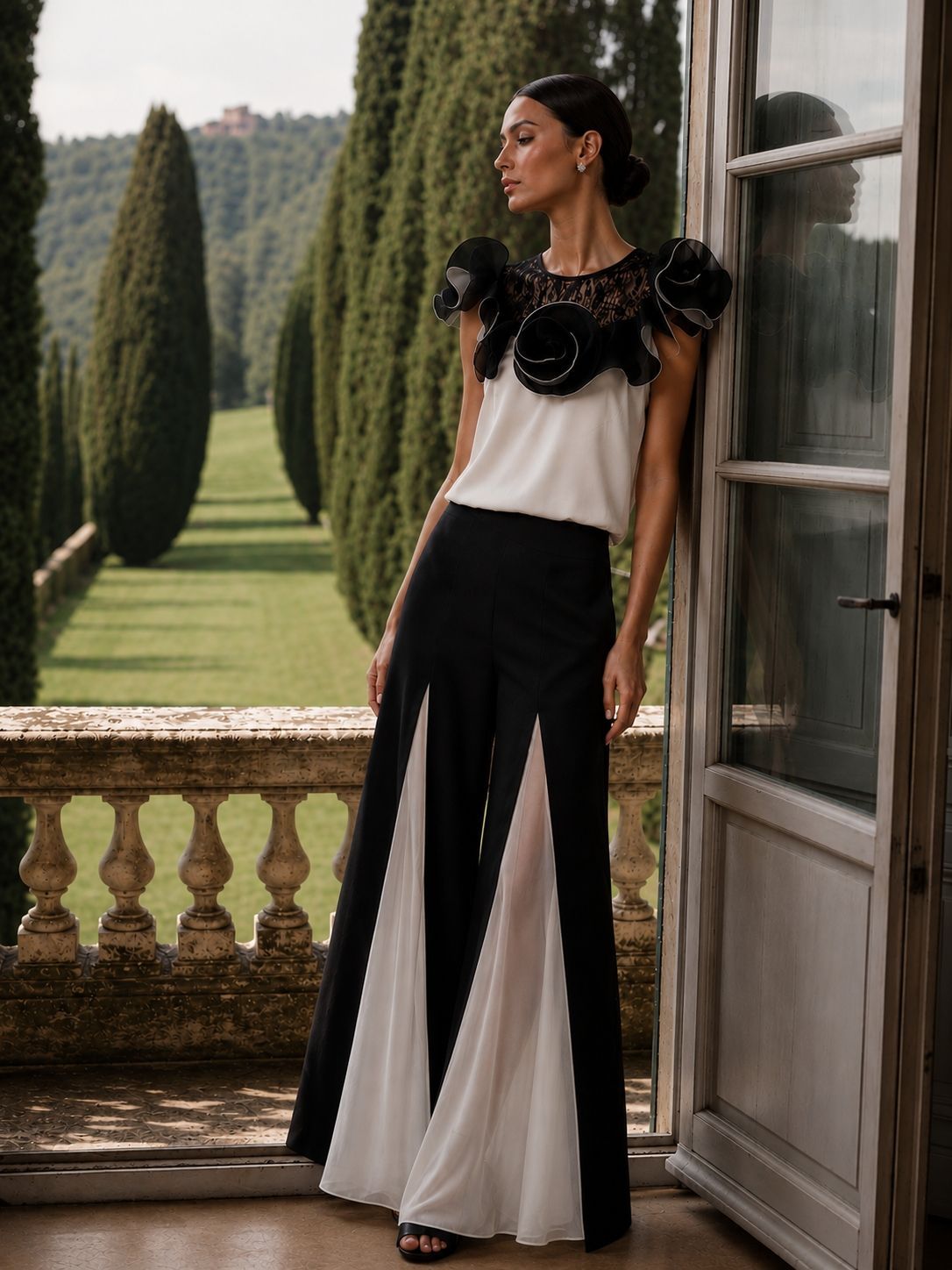 Donna con un abito bianco e nero appoggiata allo stipite di una porta su un balcone, con siepi curate alle sue spalle