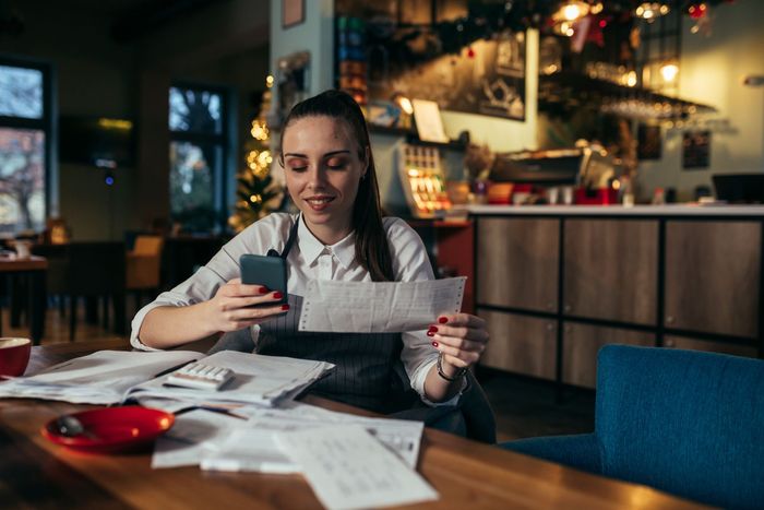 Woman reviewing documents and a phone at a table in a cozy cafe, with papers spread out nearby.