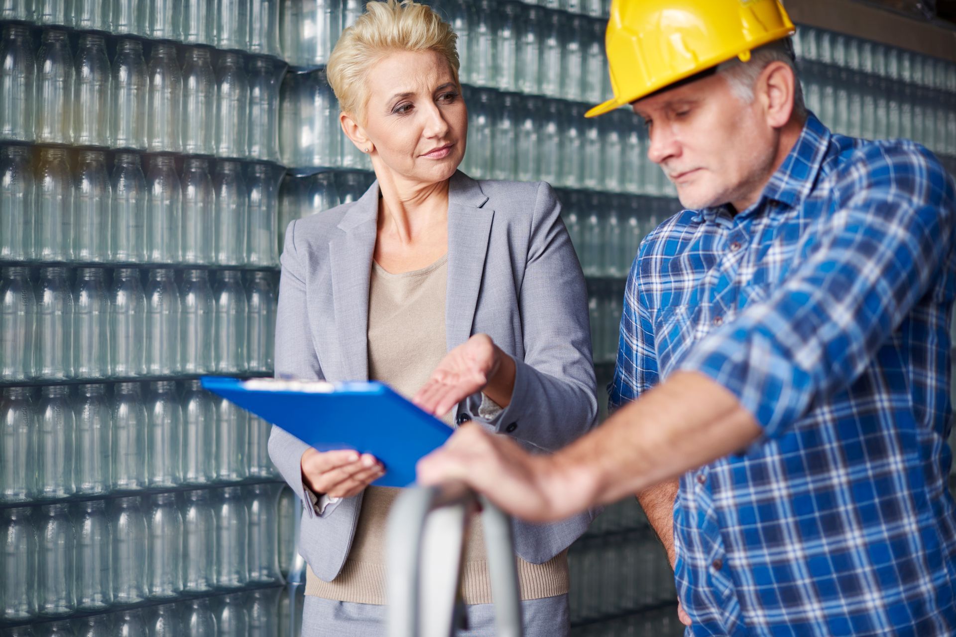 Une femme en blazer gris et un homme portant un casque de chantier jaune examinent des documents dans un entrepôt.