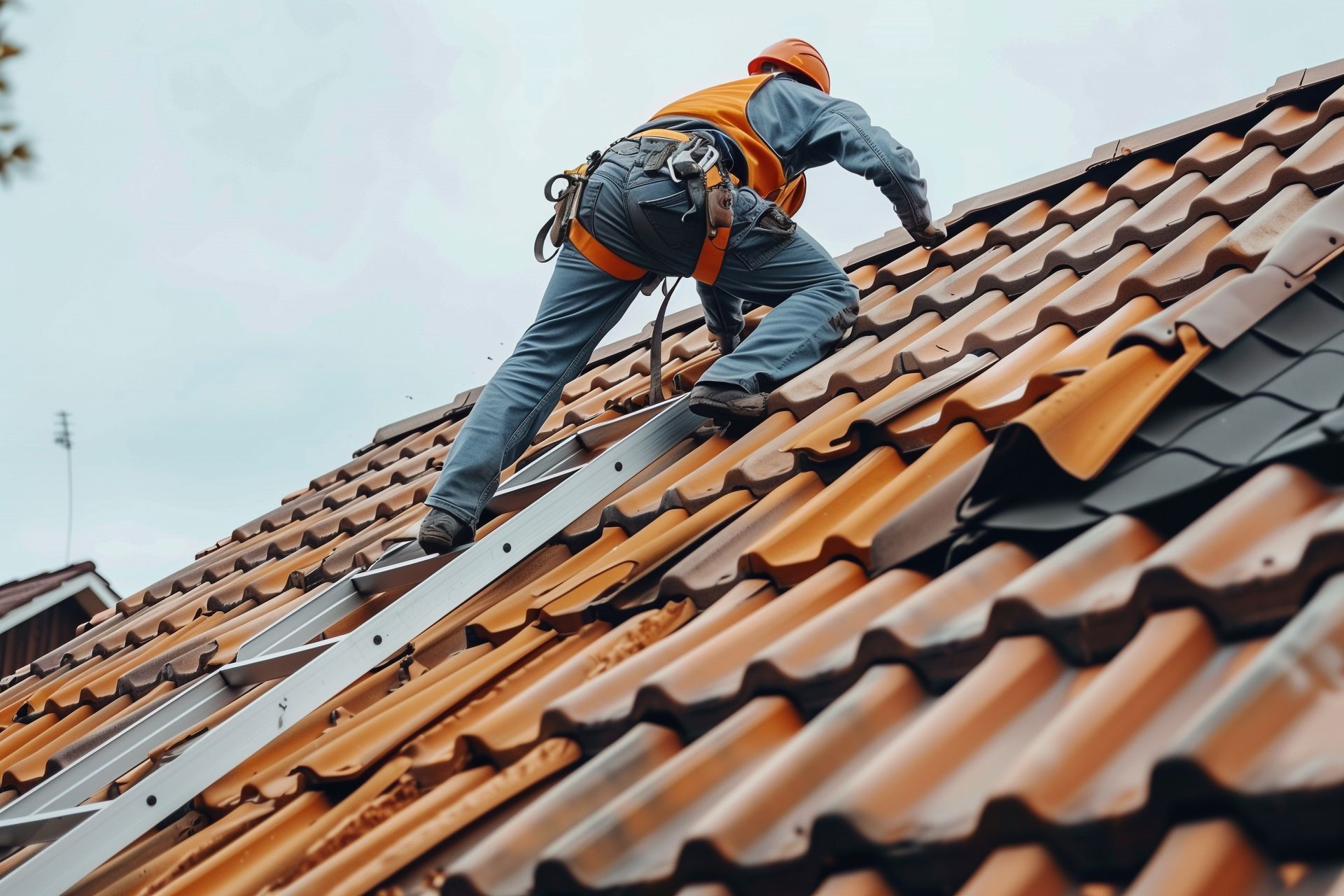 Couvreur en gilet orange et casque de chantier sur un toit de tuiles, maintenu par un harnais de sécurité.