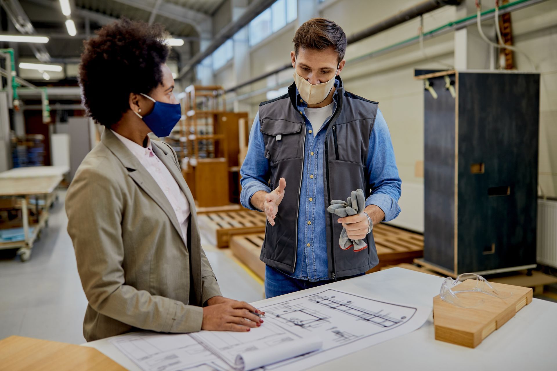 Un homme et une femme dans un atelier, examinant des plans. Tous deux portent des masques. L'homme tient un outil et fait un geste.