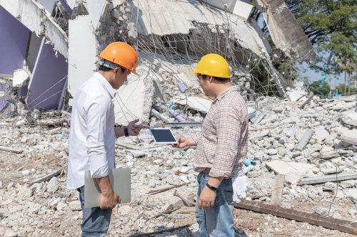 Deux personnes portant des casques de chantier examinent une tablette devant un bâtiment effondré.