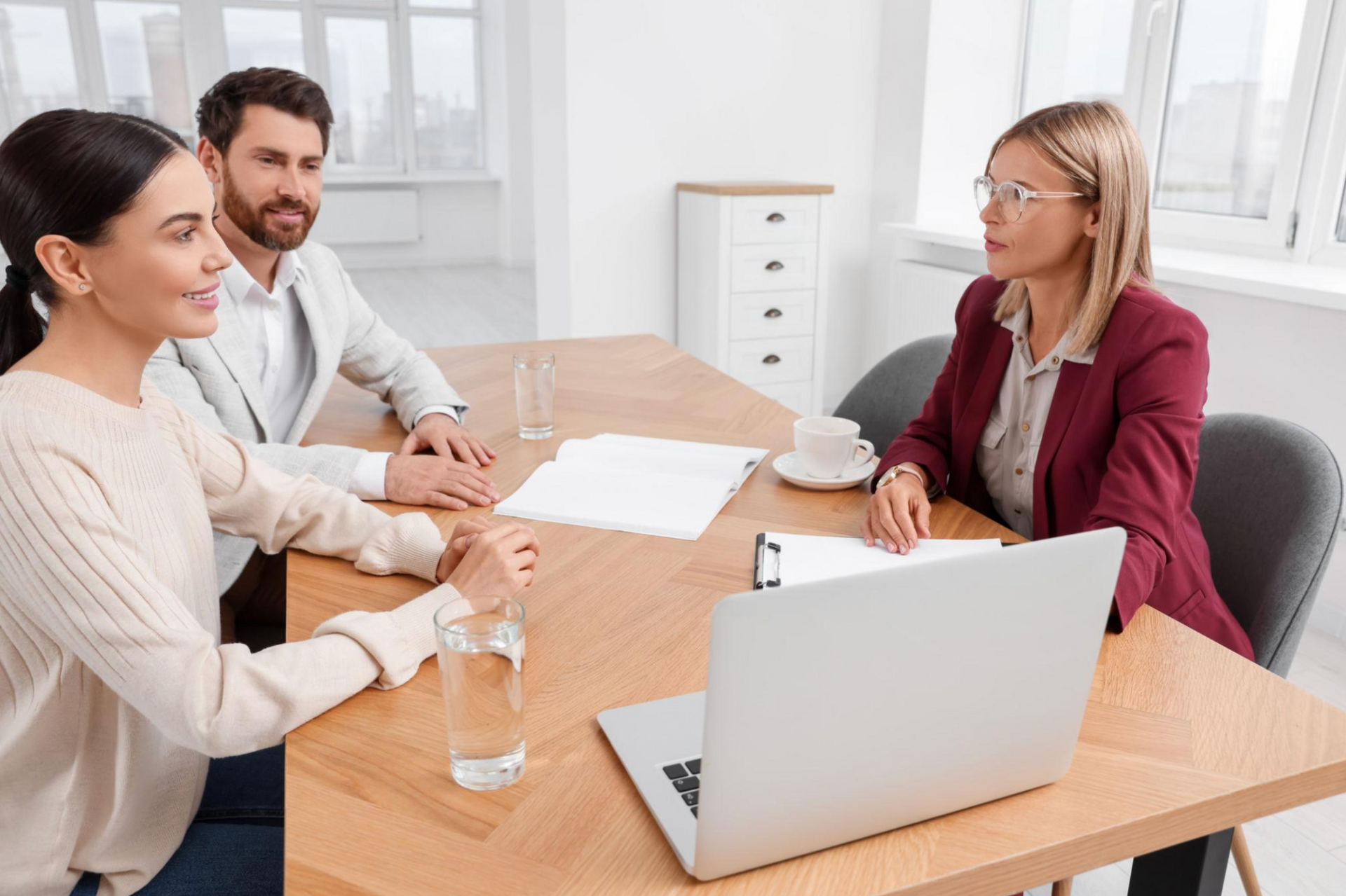 Three people in a meeting across a desk, with papers, a laptop, and a glass of water in a bright office.
