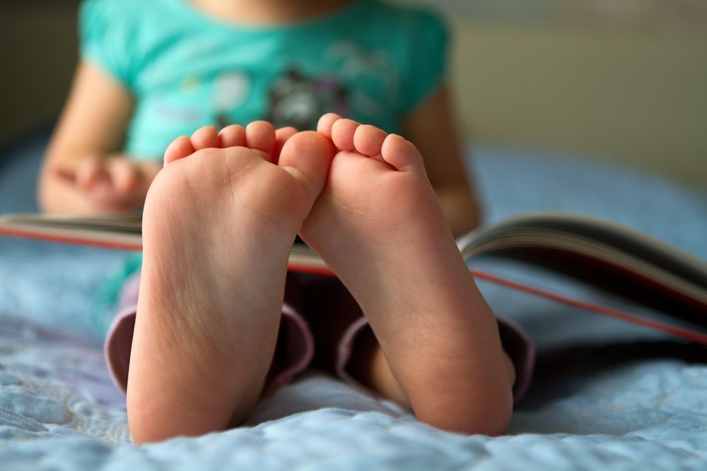 A Little Girl is Sitting on a Bed Reading a Book — Craig Hughes Podiatry in Mount Hutton, NSW