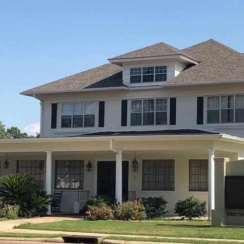 Two-story beige house with white columns, dark shutters, and a front porch under a clear blue sky