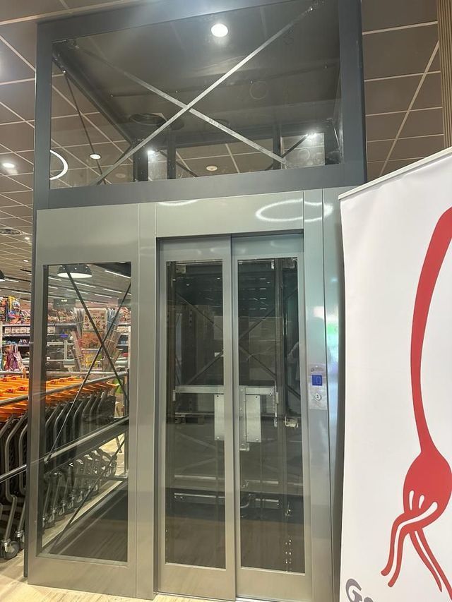 Ascensore in un supermercato con porte in vetro e struttura in acciaio inossidabile, accanto ai carrelli della spesa.
