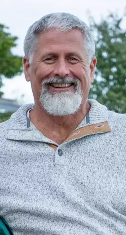 Smiling person with gray hair and a beard, wearing a gray, textured collared sweater outdoors.