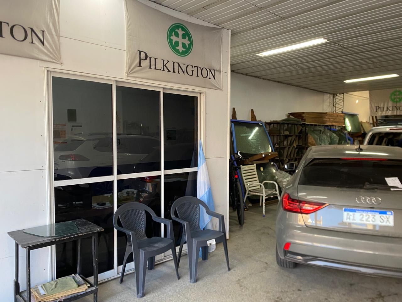 Sala de exposición de coches interior con un sedán gris aparcado junto a puertas de cristal y sillas bajo un letrero de Pillington.
