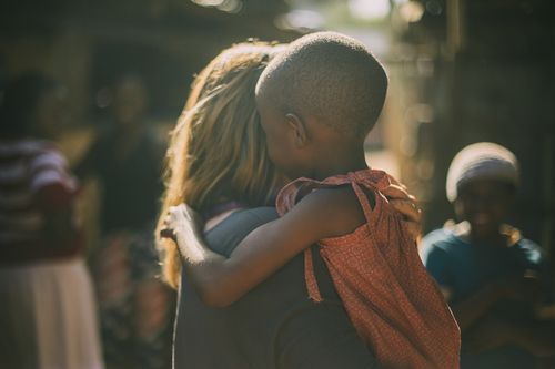 Child in pink dress hugging a person; blurred outdoor setting with other people.