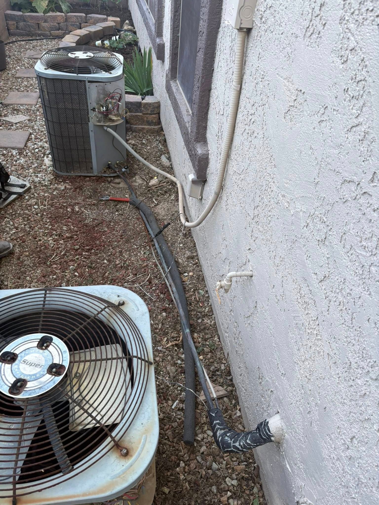 Outdoor air conditioner units beside a white wall, with hoses and gravel ground.