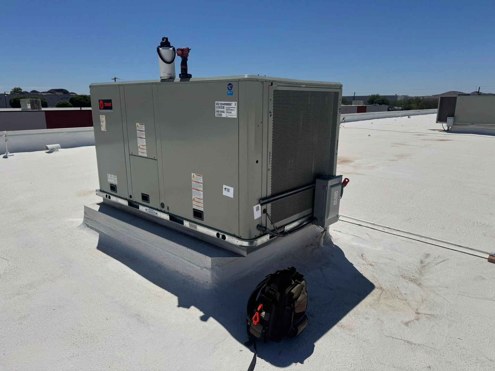 Gray rooftop HVAC unit on a white flat roof with a black tool bag in front