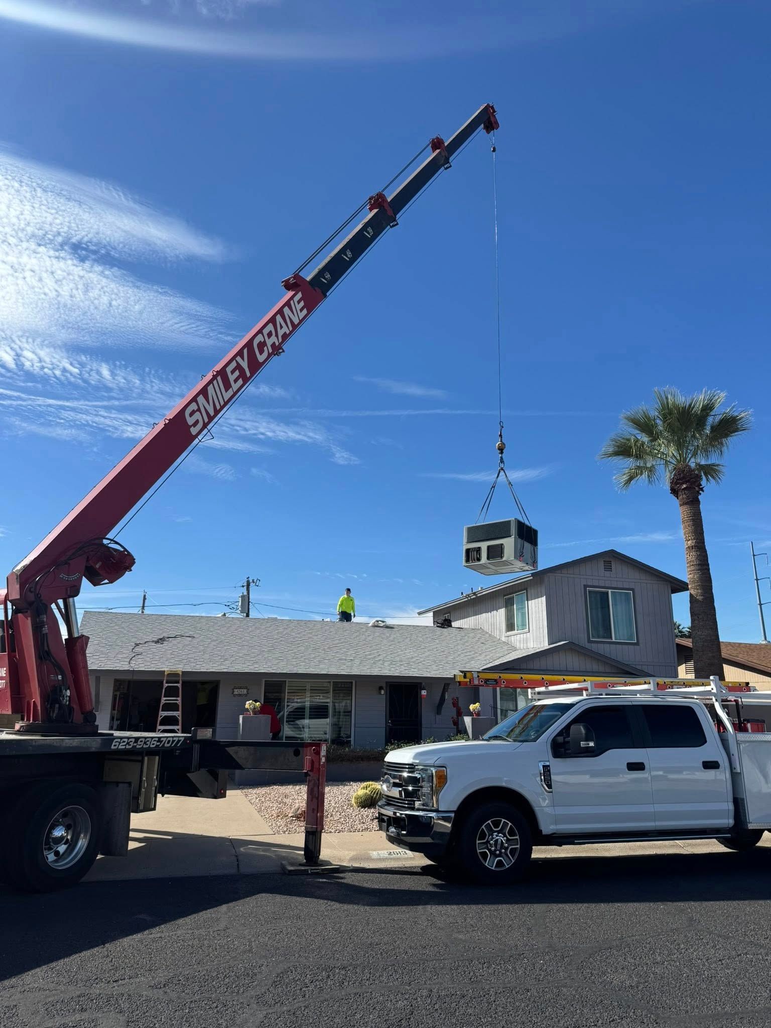 Crane lifting HVAC unit onto a rooftop at a house, with a pickup truck and palm tree nearby.