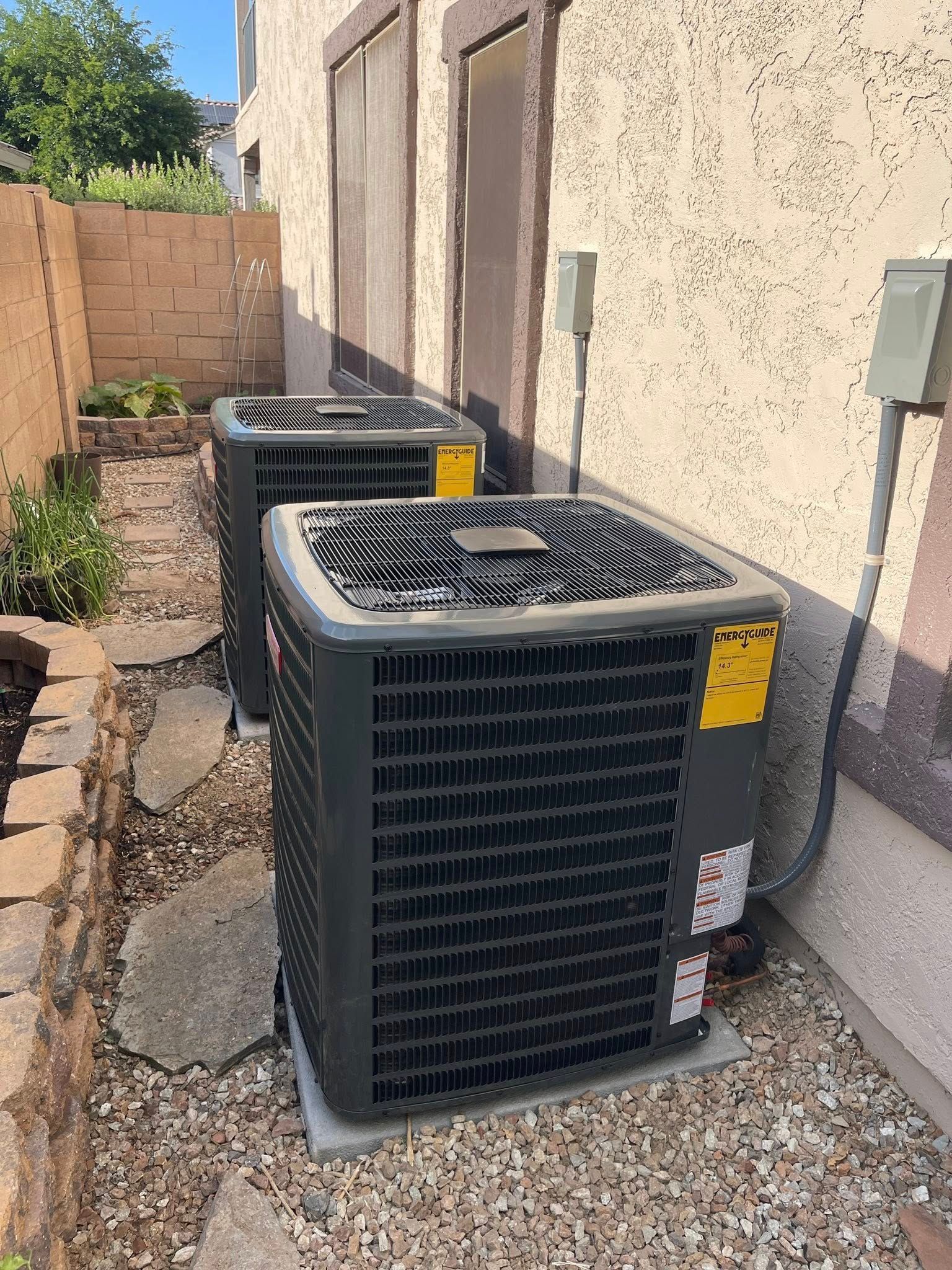 Two outdoor HVAC units beside a stucco house wall on a gravel side yard