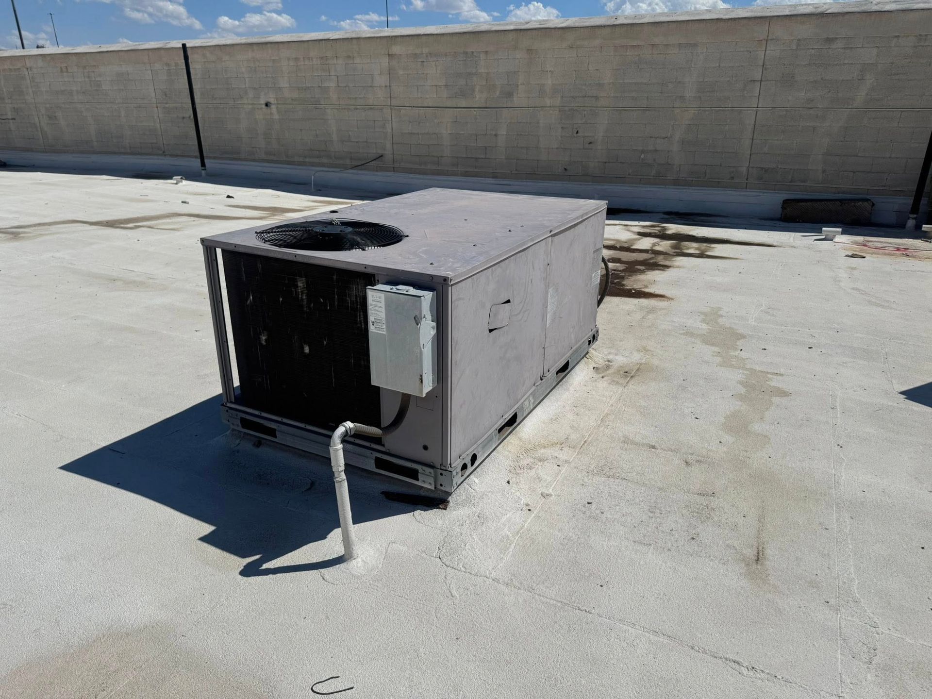 Small rooftop HVAC unit with a black front panel, duct pipe, and shadow on a concrete roof.