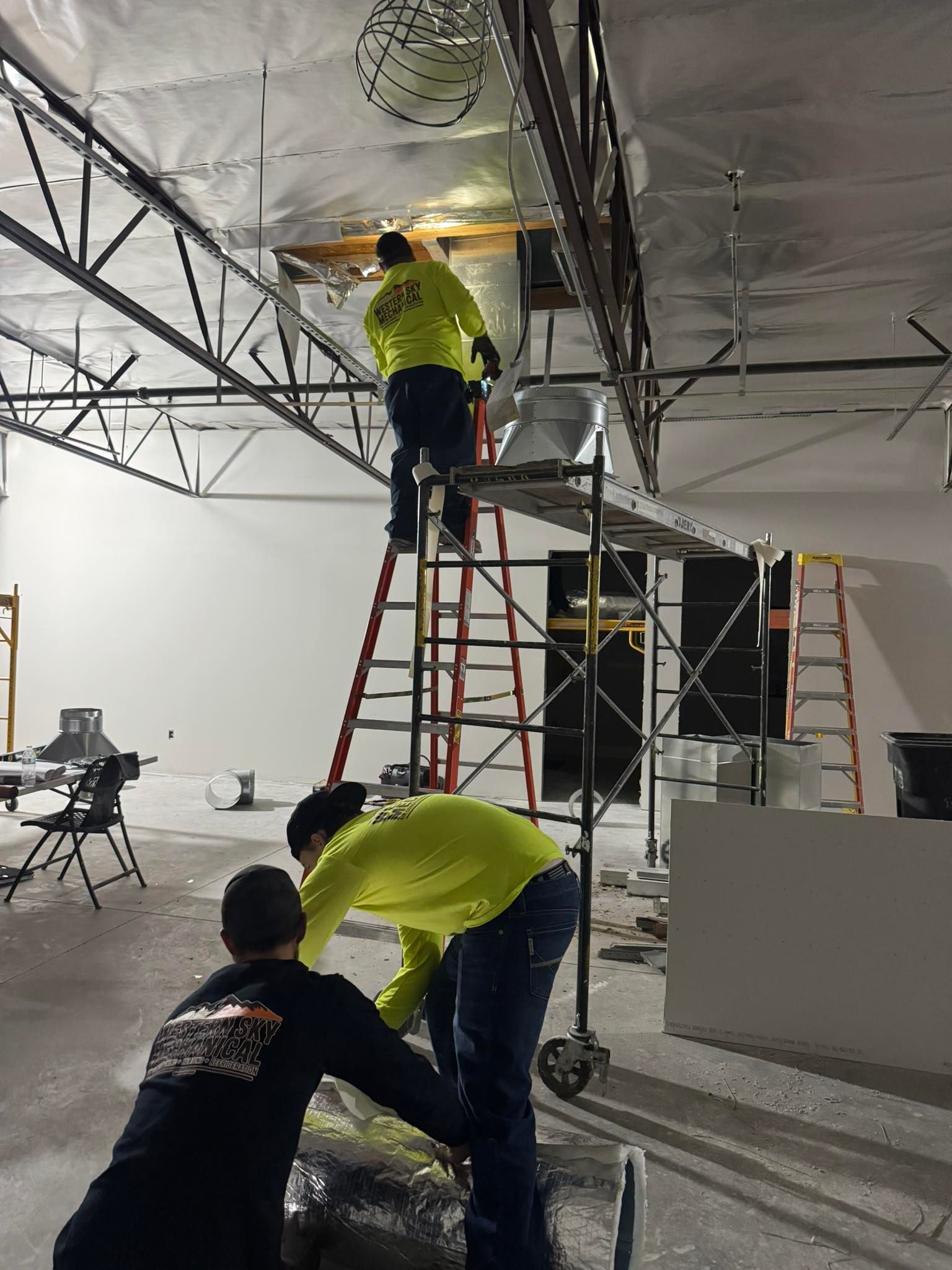 Western Sky Mechanical Workers in yellow shirts installing Hvac Duct on ladders in a large indoor space