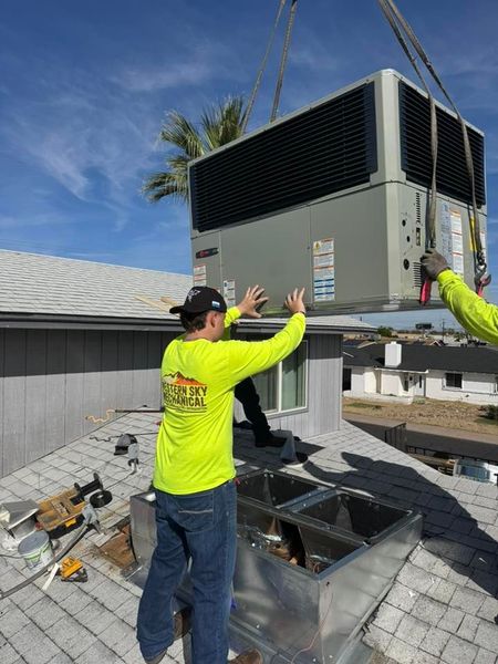 Two workers installing a large rooftop HVAC unit onto a gray shingle roof near a vent opening Phoenix AZ
