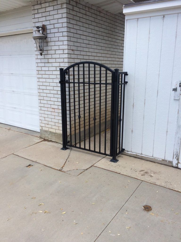Black metal gate between a white garage door and a white wooden door, set in concrete.