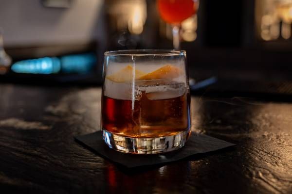 A whiskey cocktail with an orange garnish sits on a black napkin on a bar counter.