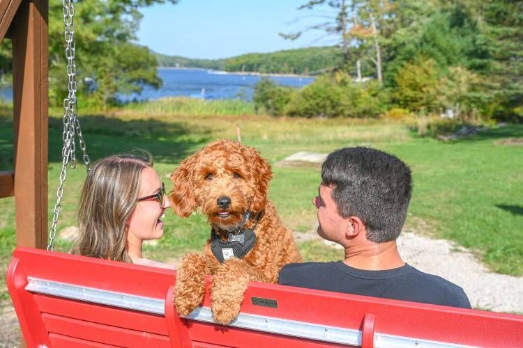 Two people on a red bench with a curly brown dog, overlooking a lake and green yard